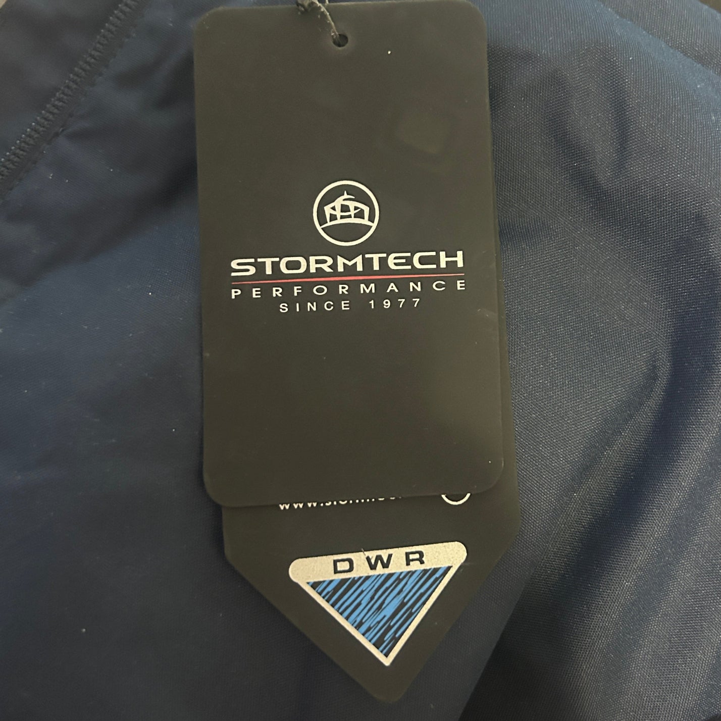 A close-up of dark navy blue outdoor apparel with a prominent Stormtech hangtag attached. The hangtag is black with white branding text and a blue and white DWR (Durable Water Repellent) sticker at the bottom.