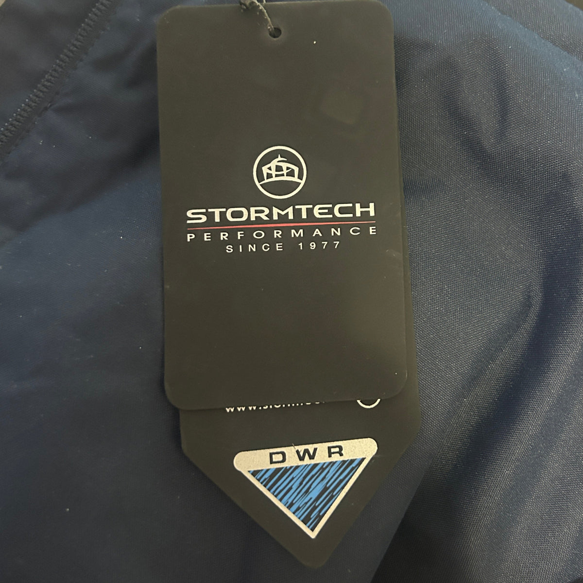 A close-up of dark navy blue outdoor apparel with a prominent Stormtech hangtag attached. The hangtag is black with white branding text and a blue and white DWR (Durable Water Repellent) sticker at the bottom.
