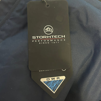 A close-up of dark navy blue outdoor apparel with a prominent Stormtech hangtag attached. The hangtag is black with white branding text and a blue and white DWR (Durable Water Repellent) sticker at the bottom.